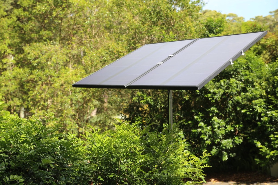 A solar panel tilted on a single pole frame standing in front of garden shrubs