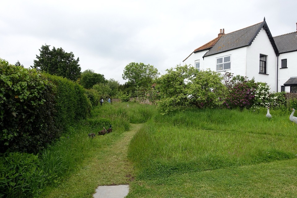 A long back garden of rough grass with a white house at the top of it