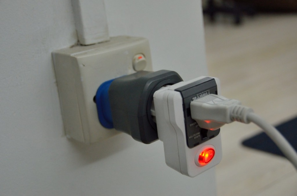 A three pin plug pushed into a white wall socket with a red indicator lit